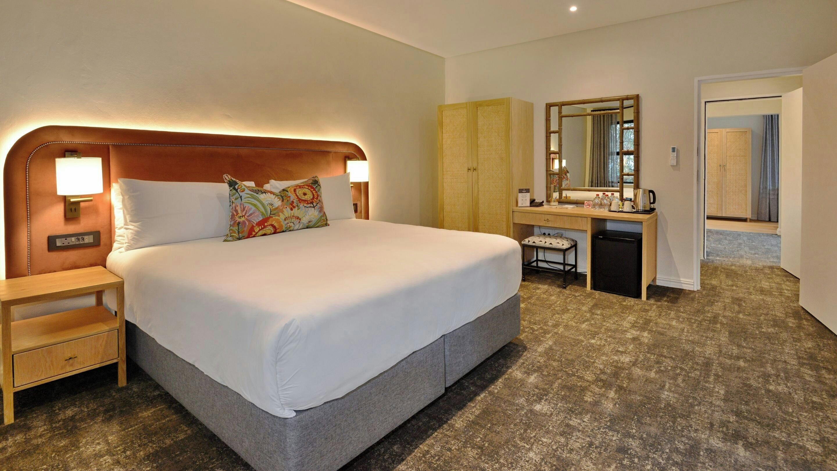 Ein Hotelzimmer mit einem großen Bett, Nachttischlampen, einem Schreibtisch mit Spiegel und einem kleinen Kühlschrank.