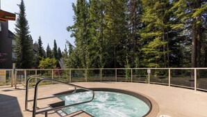 Un jacuzzi rodeado por una terraza de madera con barandilla de metal, frente a un fondo de árboles altos.