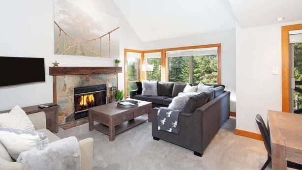 A modern living room with a fireplace, a sectional sofa, a coffee table, and large windows offering a view of greenery.