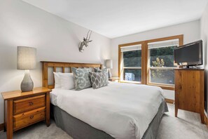 A bedroom with a bed, wooden nightstand, lamp, television, and a window with a view of greenery.