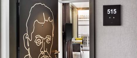 Une chambre d’hôtel moderne au design minimaliste, avec une grande œuvre d’art en forme de portrait accrochée à la porte, un téléviseur à écran plat et un balcon agrémenté d’une chaise jaune.