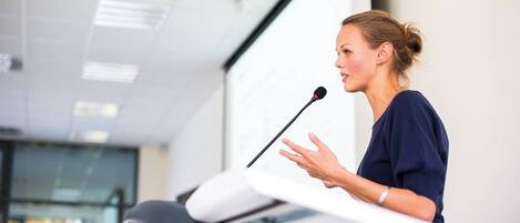 A person giving a presentation at a podium in a modern room with a glass wall and ceiling lights.