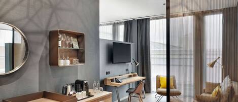 A modern hotel room with a wooden desk, a flat-screen TV, a sofa, and a large window with curtains.