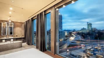 A modern hotel room with a large window, a bathtub, and a vanity with a mirror.