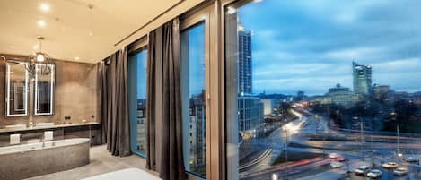 A modern hotel room with a large window, a bathtub, and a vanity with a mirror.