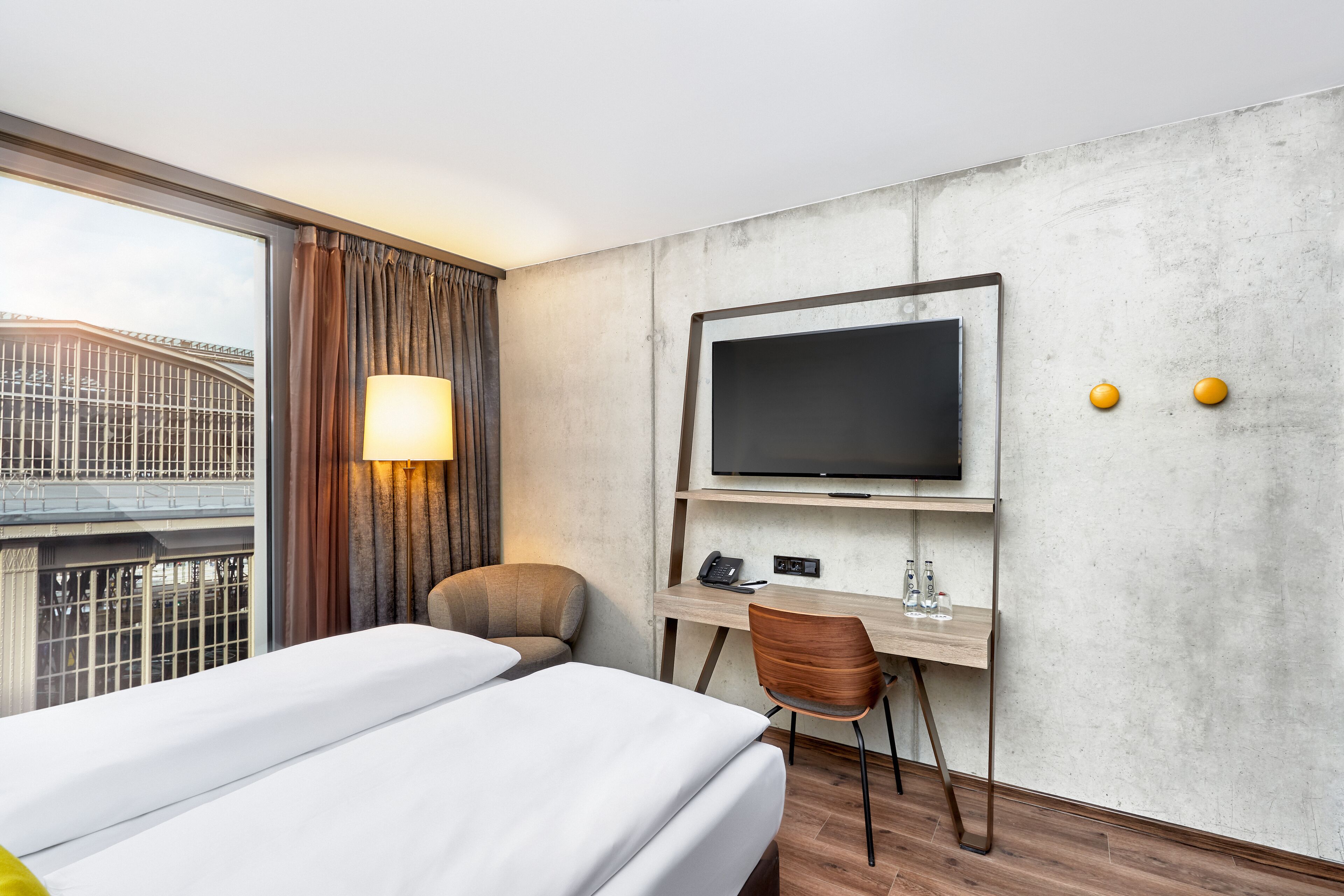 A hotel room with a bed, a desk with a TV, a chair, and a window with curtains.