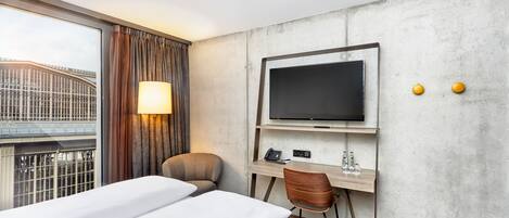A hotel room with a bed, a desk with a TV, a chair, and a window with curtains.