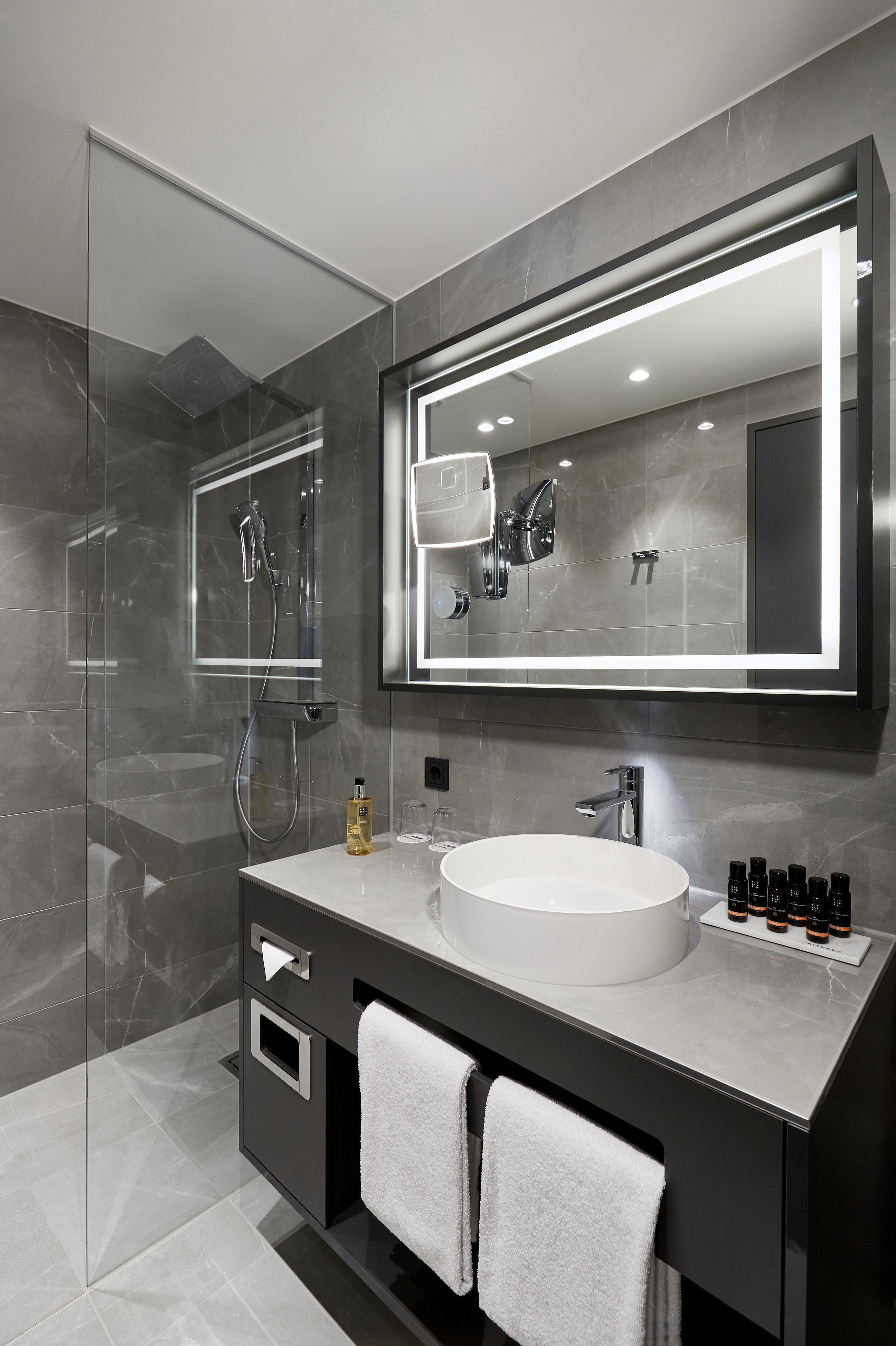 A modern bathroom with a glass shower enclosure, a large mirror, a round sink, and a vanity with a towel rack.