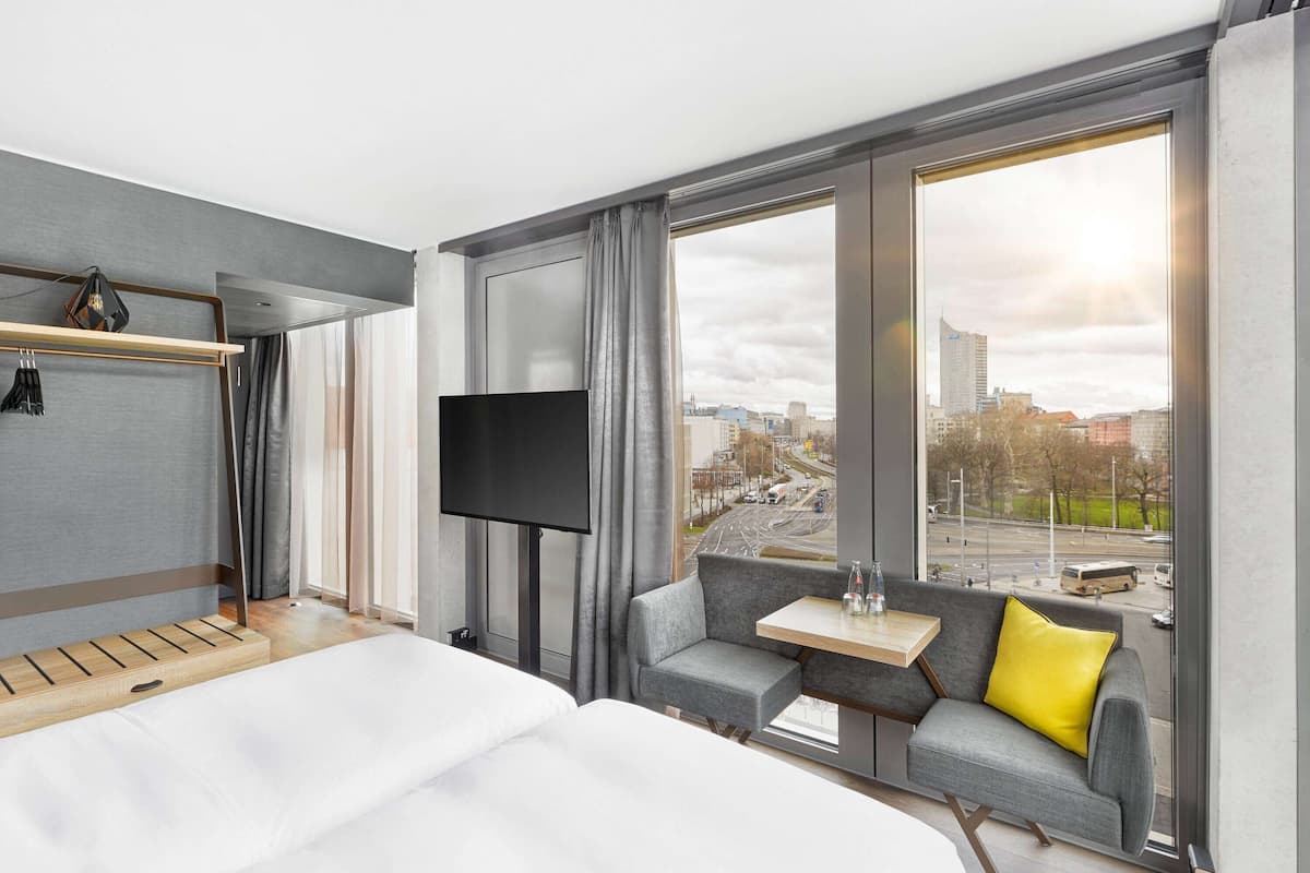 Une chambre d’hôtel avec un lit, une télévision, une petite table, un canapé et une grande fenêtre donnant sur la ville.