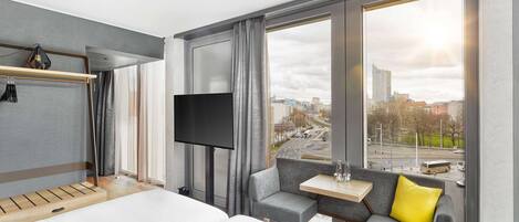 Habitación de hotel con cama, televisor, una mesita, sofá y un amplio ventanal con vistas a la ciudad.