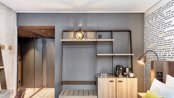 Ein modernes Hotelzimmer mit einem Bett, einer hölzernen Bank, einem Kleiderschrank und einem Schreibtisch mit Kaffeemaschine.