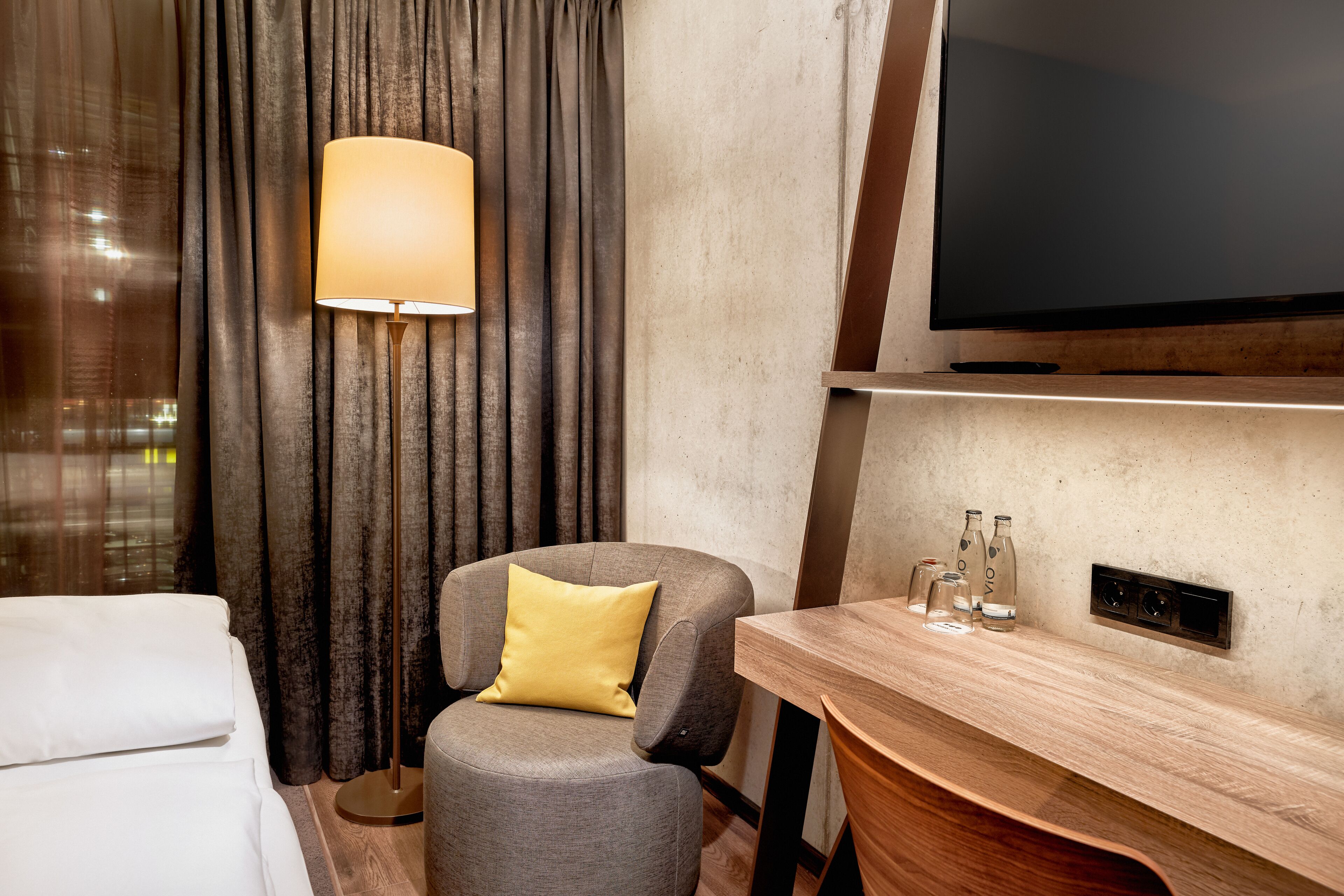 Ein Hotelzimmer mit einem Bett, einem grauen Sessel mit gelbem Kissen, einer Stehlampe, einem Holfnachtkästchen mit Flaschen und einem an der Wand befestigten Flachbildfernseher.