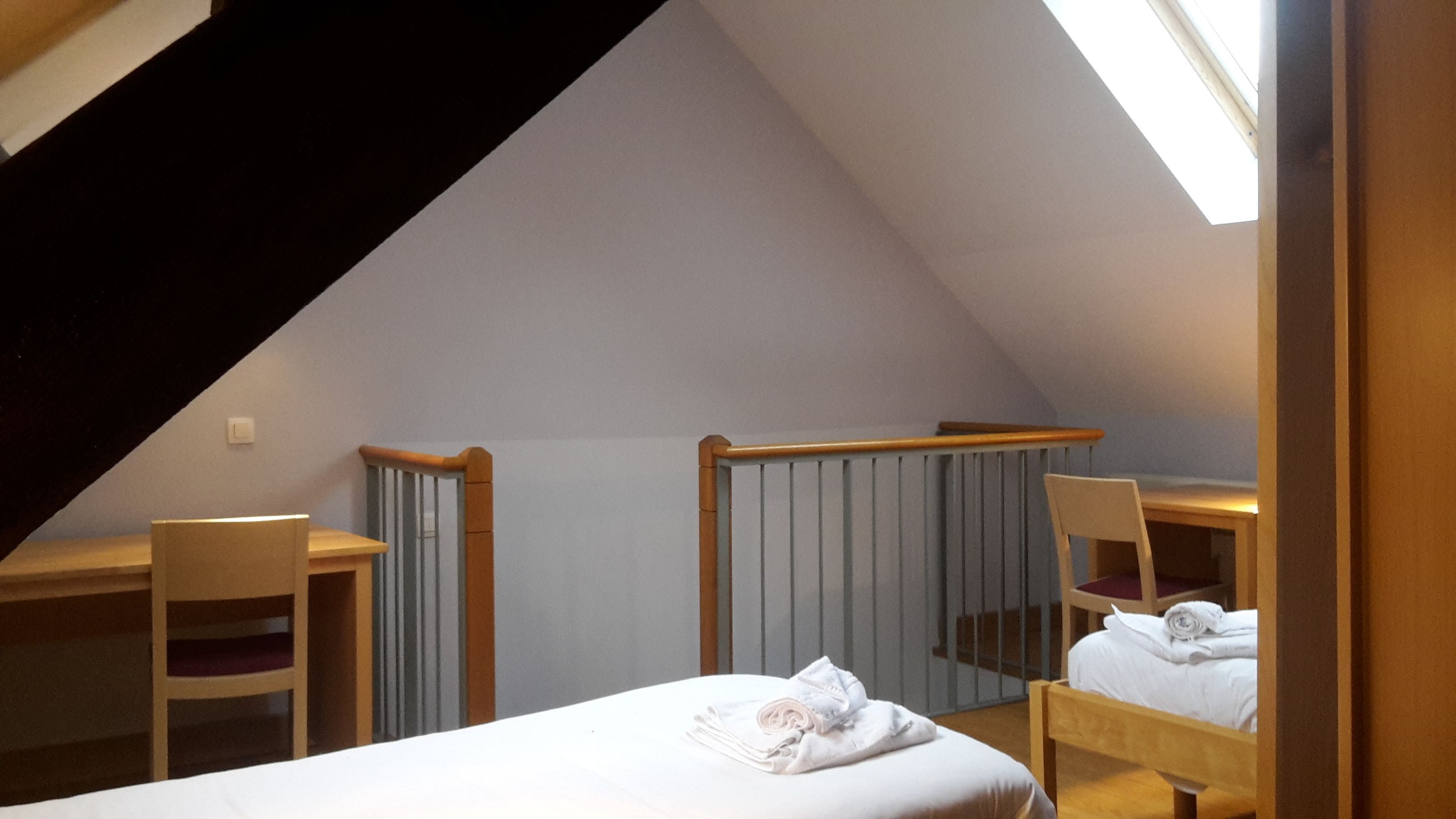 A small, triangular attic bedroom with a sloped ceiling, a wooden desk, two chairs, and a bed with neatly folded towels.