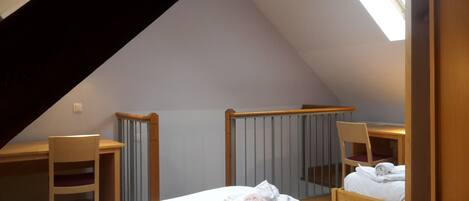 A small, triangular attic bedroom with a sloped ceiling, a wooden desk, two chairs, and a bed with neatly folded towels.