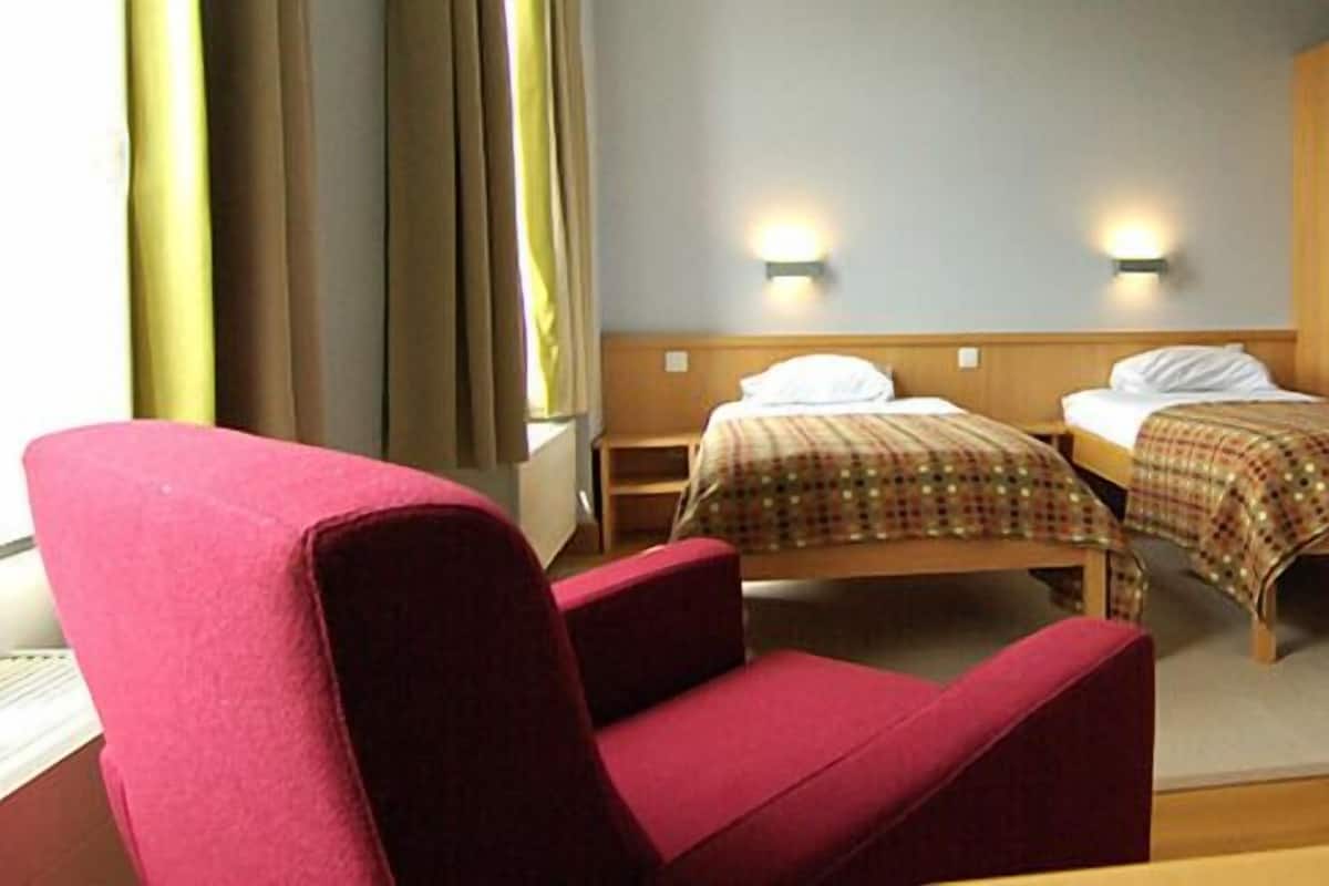 A hotel room with two beds, a red armchair, and a wooden wardrobe.