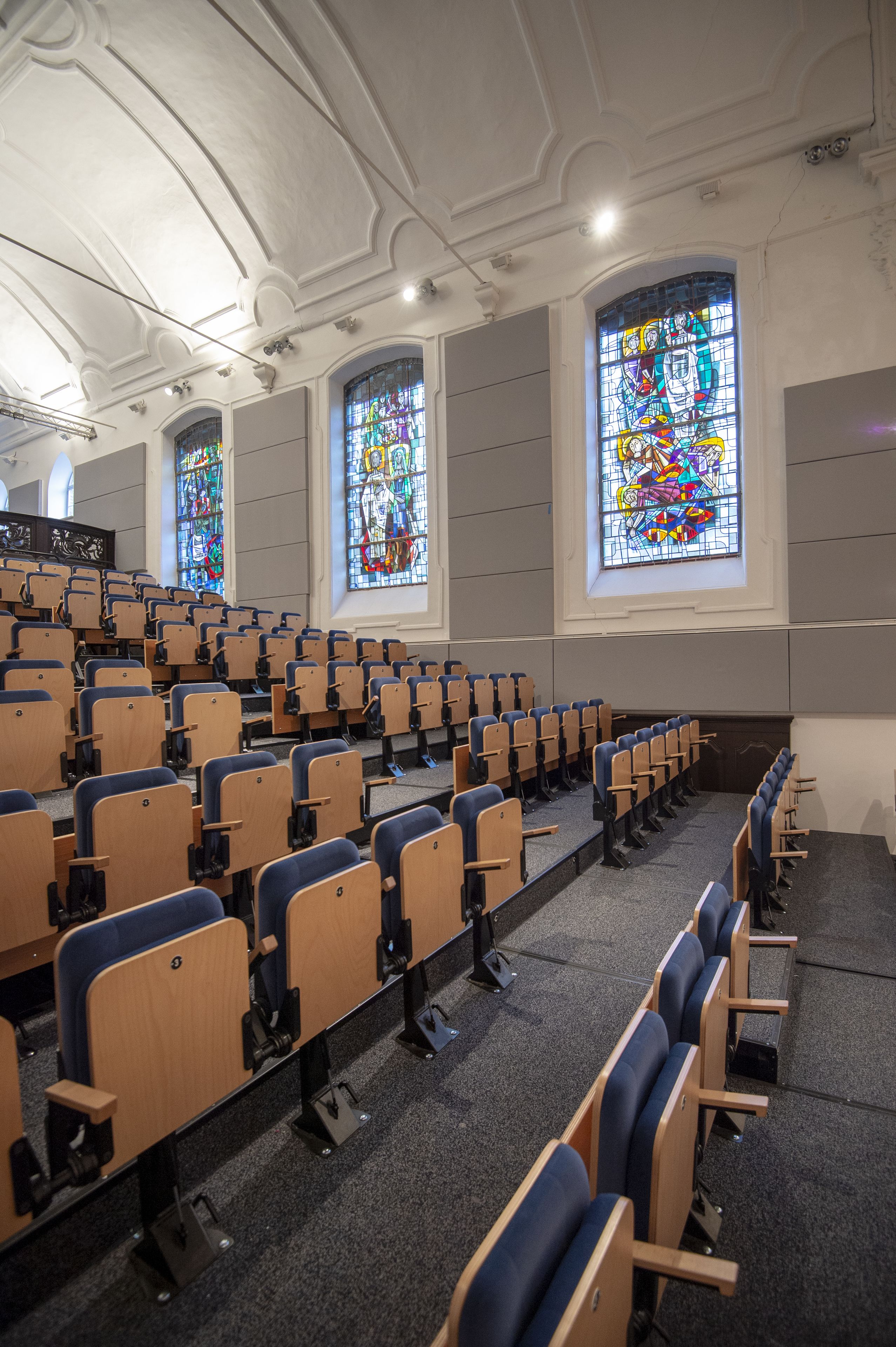 Un auditorio con filas de sillas de madera y grandes ventanales de vitral.