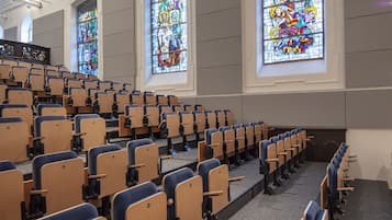 Un auditorio con filas de sillas de madera y grandes ventanales de vitral.