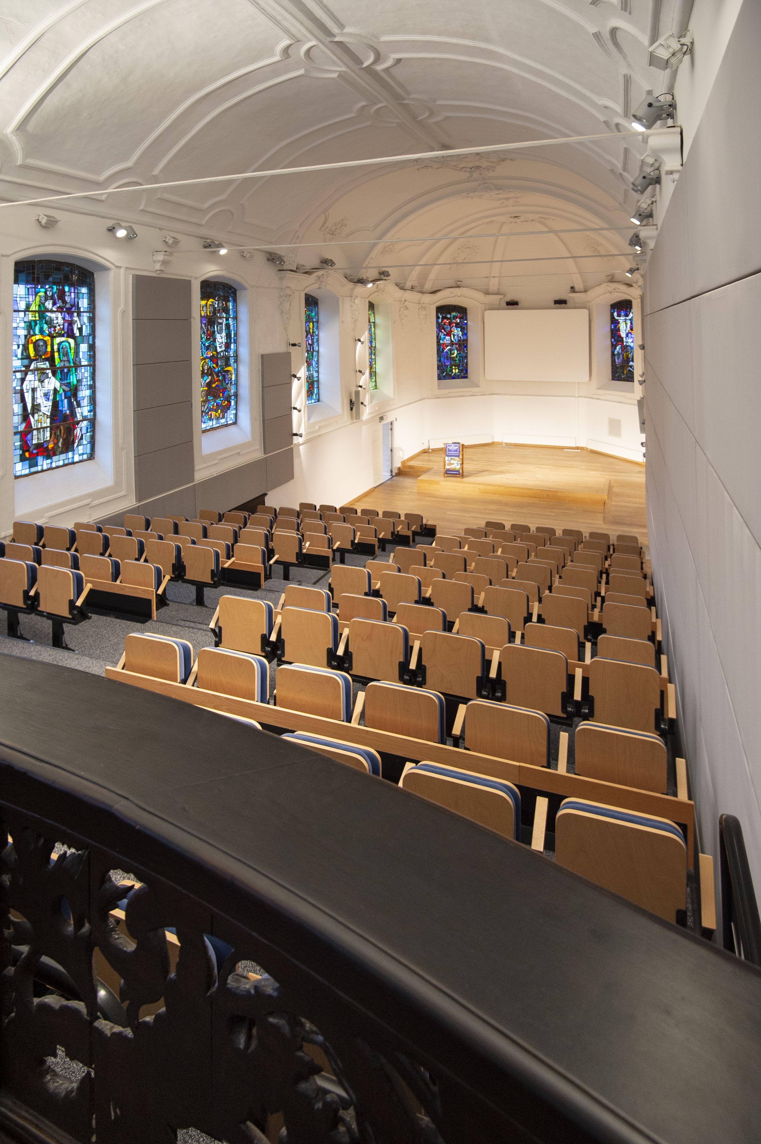 An auditorium with rows of wooden chairs and stained glass windows.