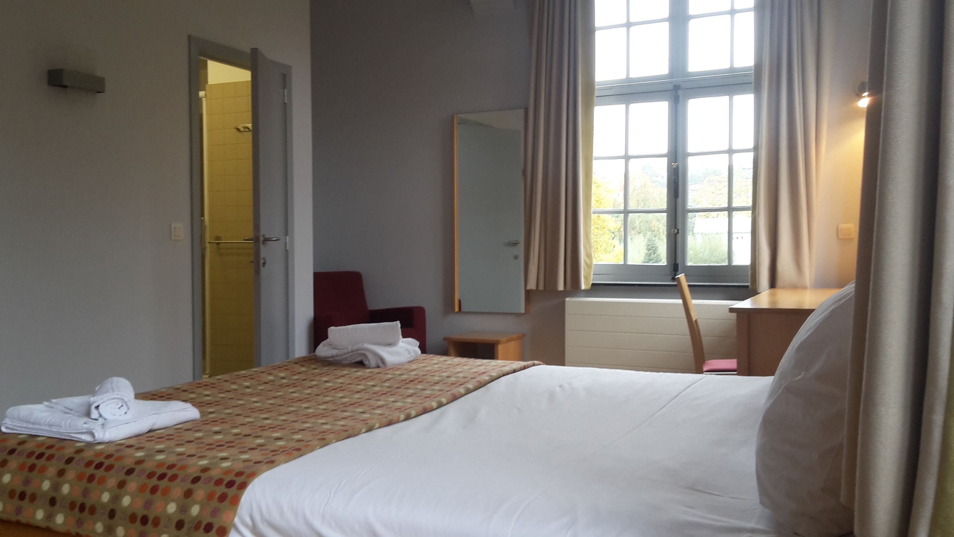 A hotel room with a bed, a bedside table, a chair, a mirror, and a window with curtains.