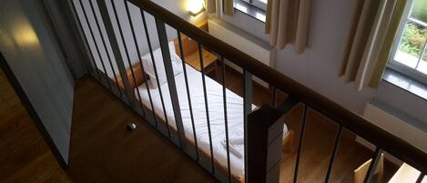 Un dormitorio con piso de madera, una cama con ropa blanca, una mesita de noche y una ventana con cortinas.