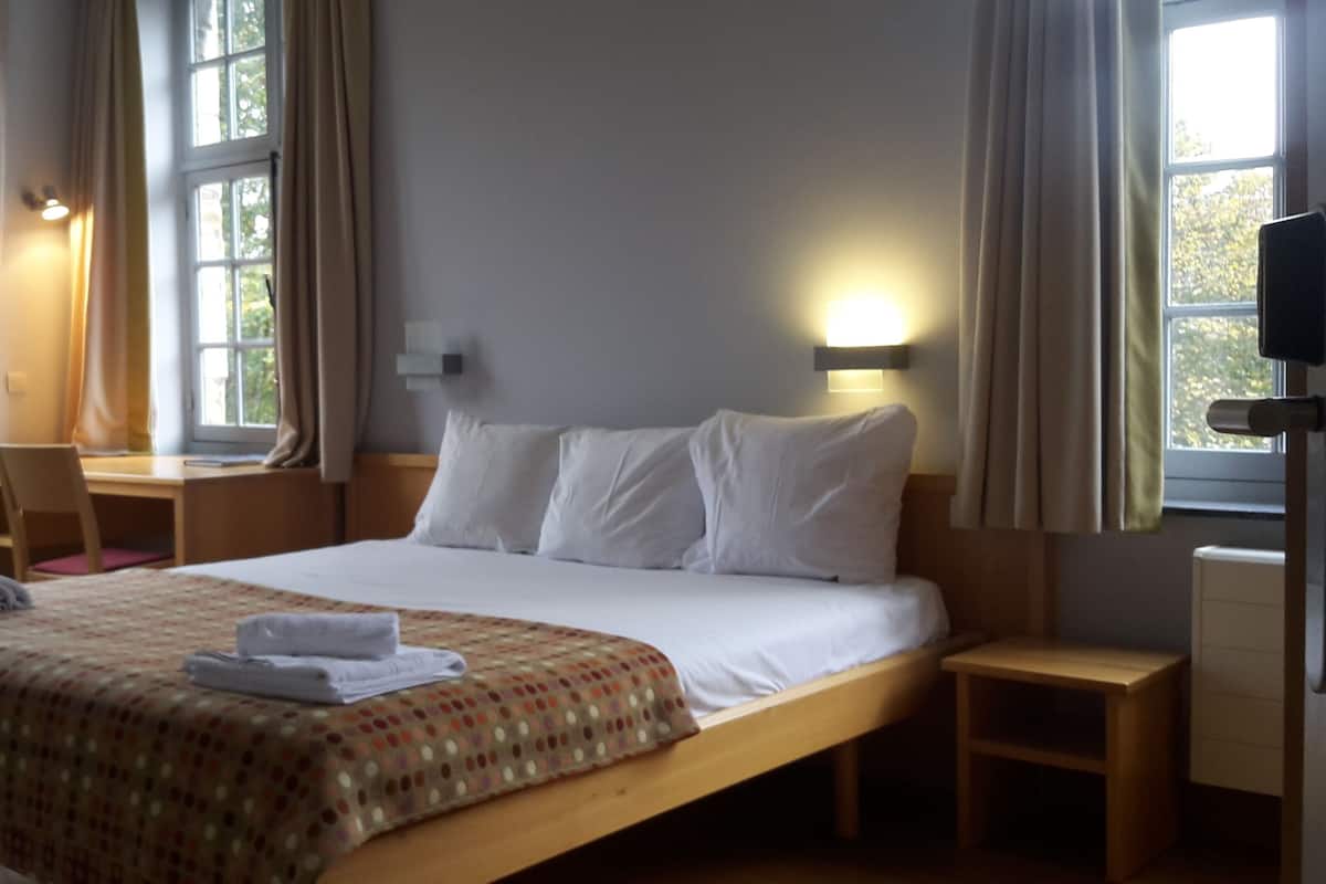 A hotel room with a large bed, two bedside tables, a desk, and a chair. There are two windows with curtains, and a wall-mounted TV.