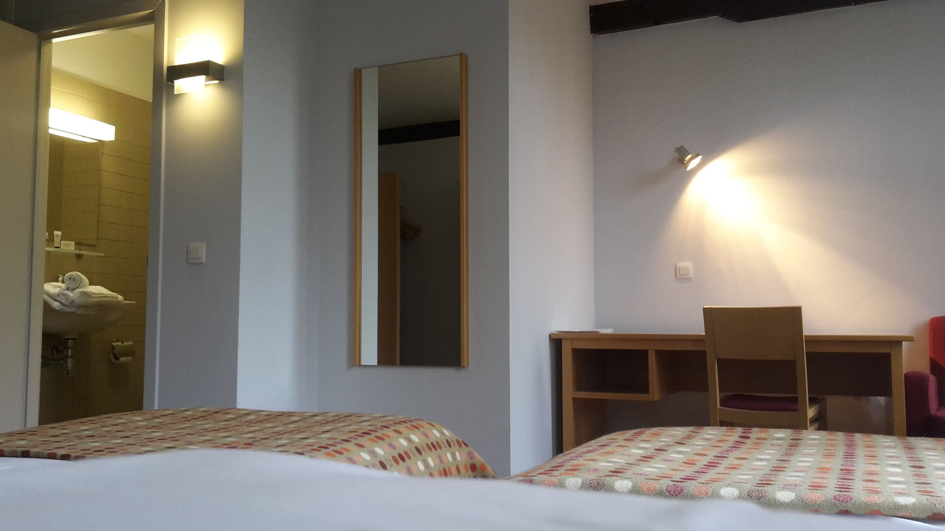 A hotel room with two beds, a desk, a chair, a mirror, and a bathroom with a sink and towel rack.