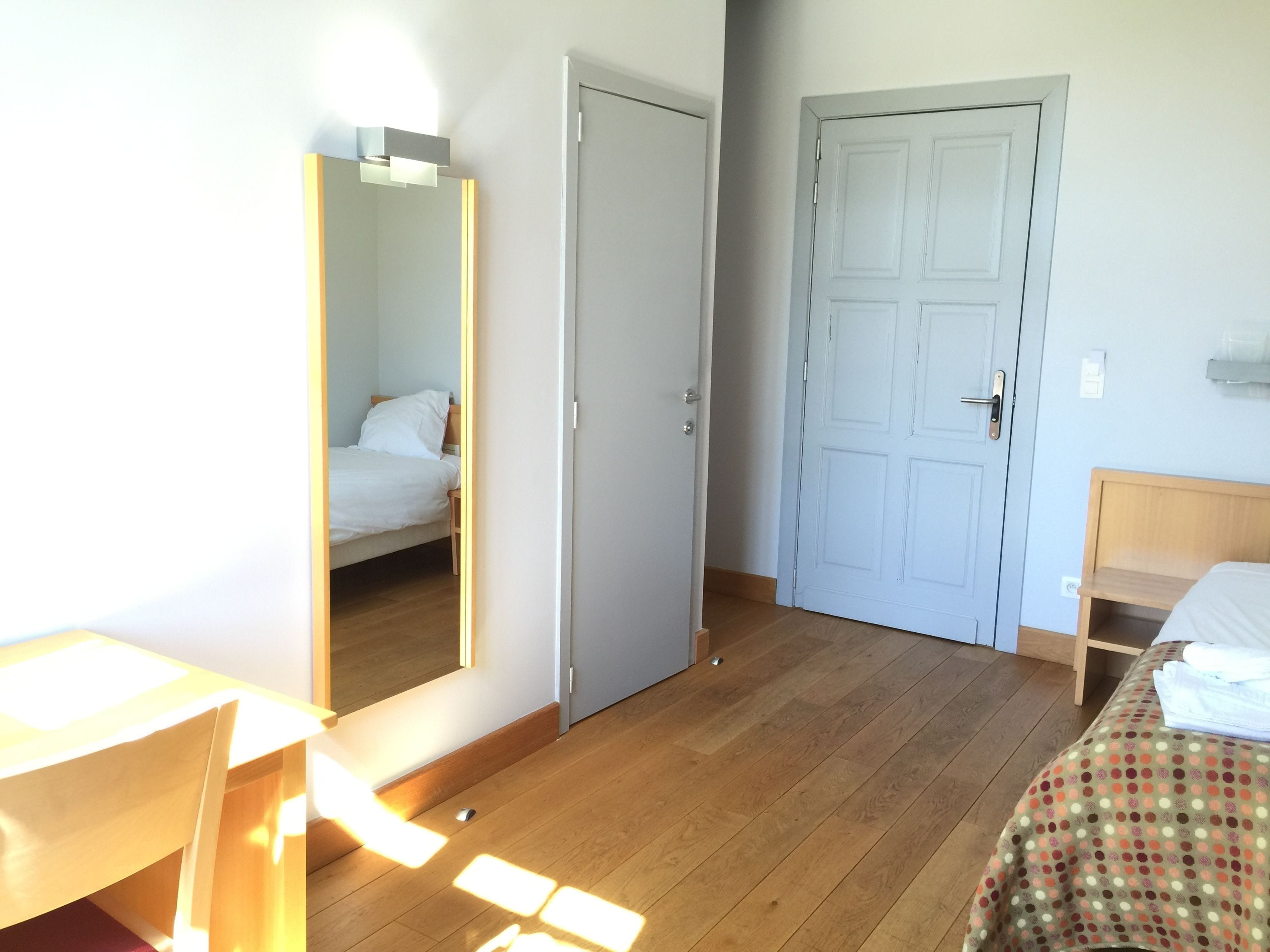 Una habitación con piso de madera, una cama con cubrecama blanco, un escritorio con silla y una puerta.