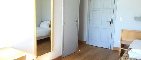 Una habitación con piso de madera, una cama con cubrecama blanco, un escritorio con silla y una puerta.