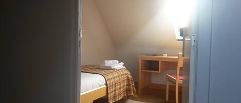 A small, dimly lit room with a single bed, a desk, and a chair.