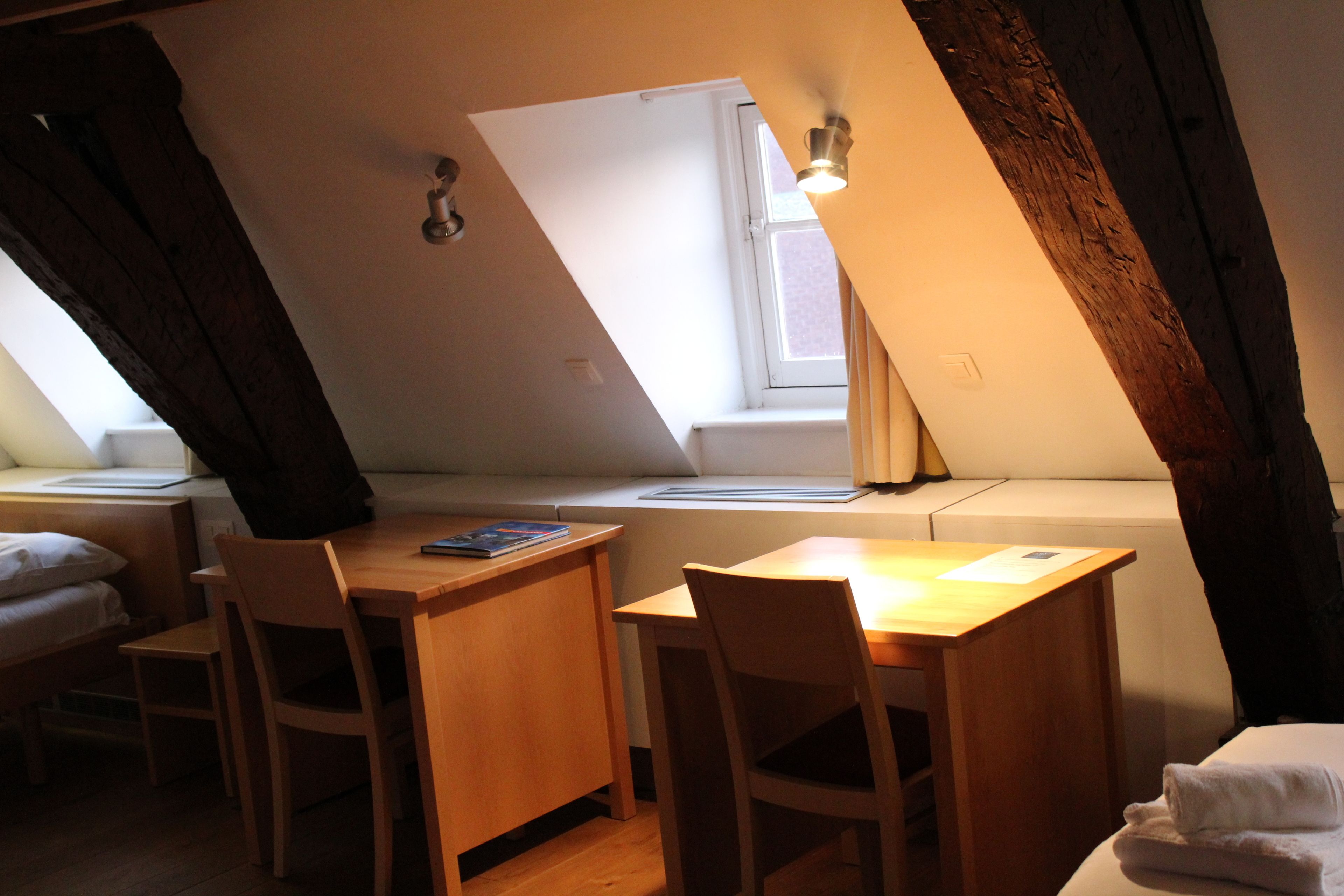 A room with a sloped ceiling, a wooden desk with two chairs, a window with a view, and a bed with a white blanket.