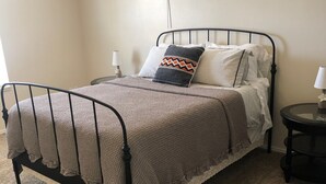 A neatly made bed with a metal frame, a patterned throw blanket, and a decorative pillow. There is a small round table with a lamp beside the bed, and a window with blinds in the background. - Your Home Away From Home in Escalante (Escalante)