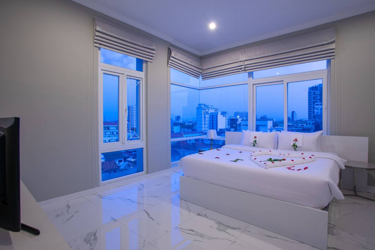 A modern bedroom with a large bed, a TV, and a city view through large windows.