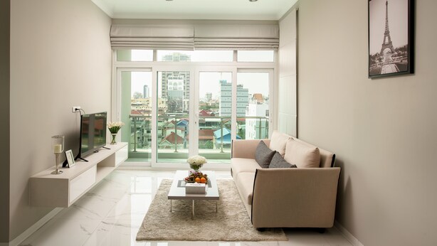 A modern living room with a sofa, a coffee table with fruit and flowers, a TV on a stand, and a city view through large windows. - SSB Residence (Phnom Penh)