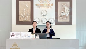 A reception desk with two individuals standing behind it. There are clocks on the wall displaying different times, and a sign with a no-smoking symbol. - SSB Residence (Phnom Penh)