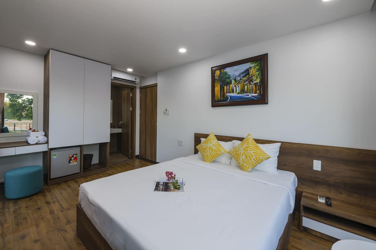 A hotel room with a bed, a desk, a chair, a small fridge, and a painting on the wall.