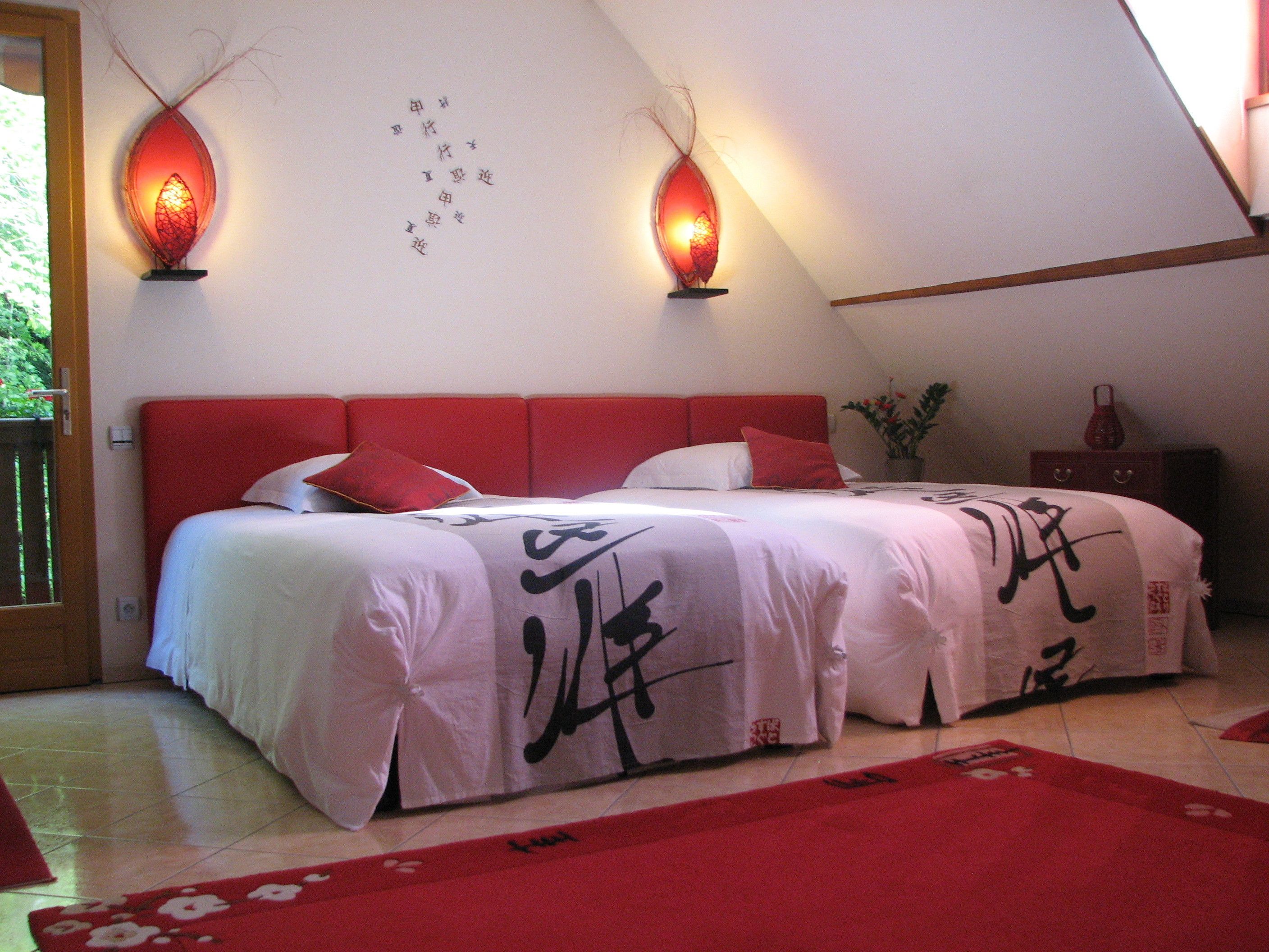 Ein Schlafzimmer mit einem roten Bettrahmen, einem Bett mit weißer Bettwäsche und roten Kissen, zwei roten Wandlampen und einer Wand mit chinesischen Schriftzeichen.