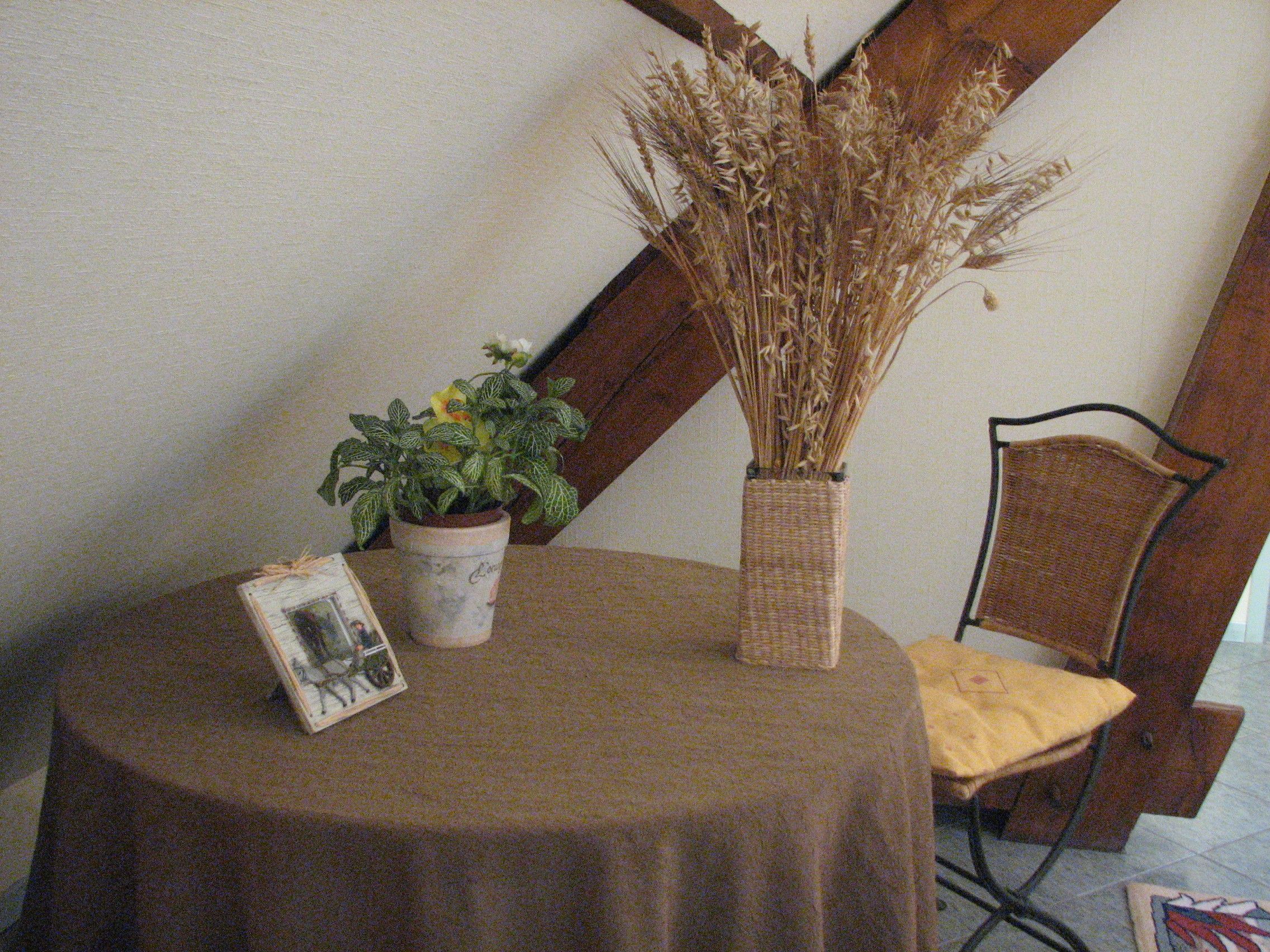 Una mesa redonda con mantel marrón, una planta en maceta, un jarrón con flores secas y una foto enmarco.