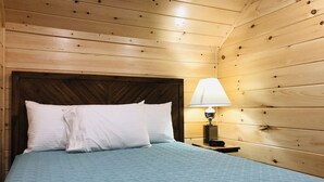Une pièce aux murs lambrissés, avec un lit, des oreillers blancs, une couvre-lit matelassée bleue, une table de chevet avec une lampe et une horloge numérique affichant 12h46.