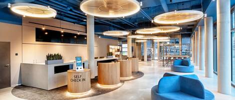 A modern reception area with a self-check-in station, large circular ceiling lights, and a blue seating area.