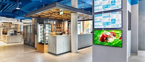Eine moderne Hotelhalle mit einer zentralen Rezeption, einer digitalen Speisekarte und einem großen Bildschirm, der Speisen zeigt.