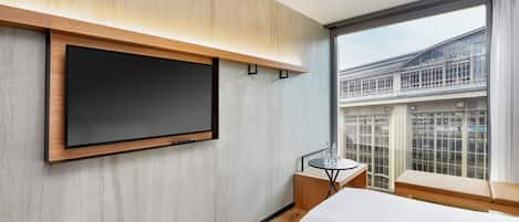 A modern hotel room with a large TV mounted on the wall, a small table with a glass top, and a large window offering a view of the city.