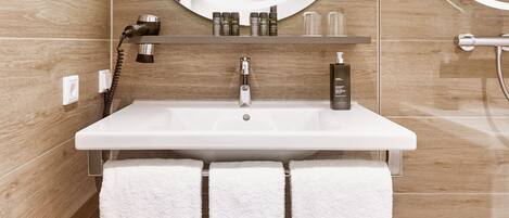 A bathroom with a white sink, two white towels, a round mirror with lights, and a shower area.