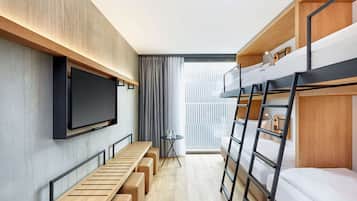 Een kleine hotelkamer met stapelbedden, een aan de muur bevestigde televisie, een klein tafeltje met een stoel en een bankje langs de muur.