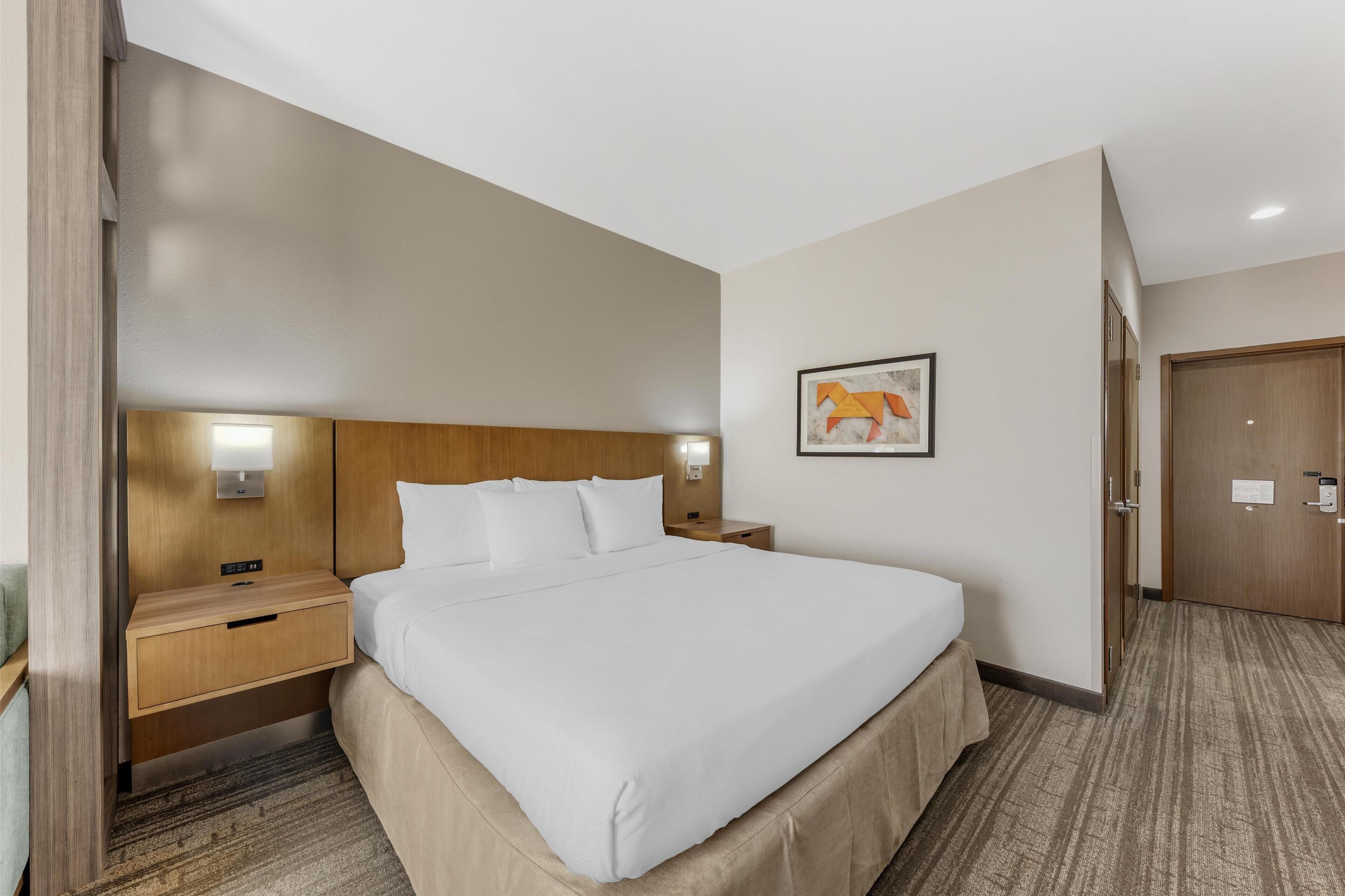 A hotel room with a large bed, wooden headboard, bedside tables with lamps, a framed picture on the wall, and a door to the right.