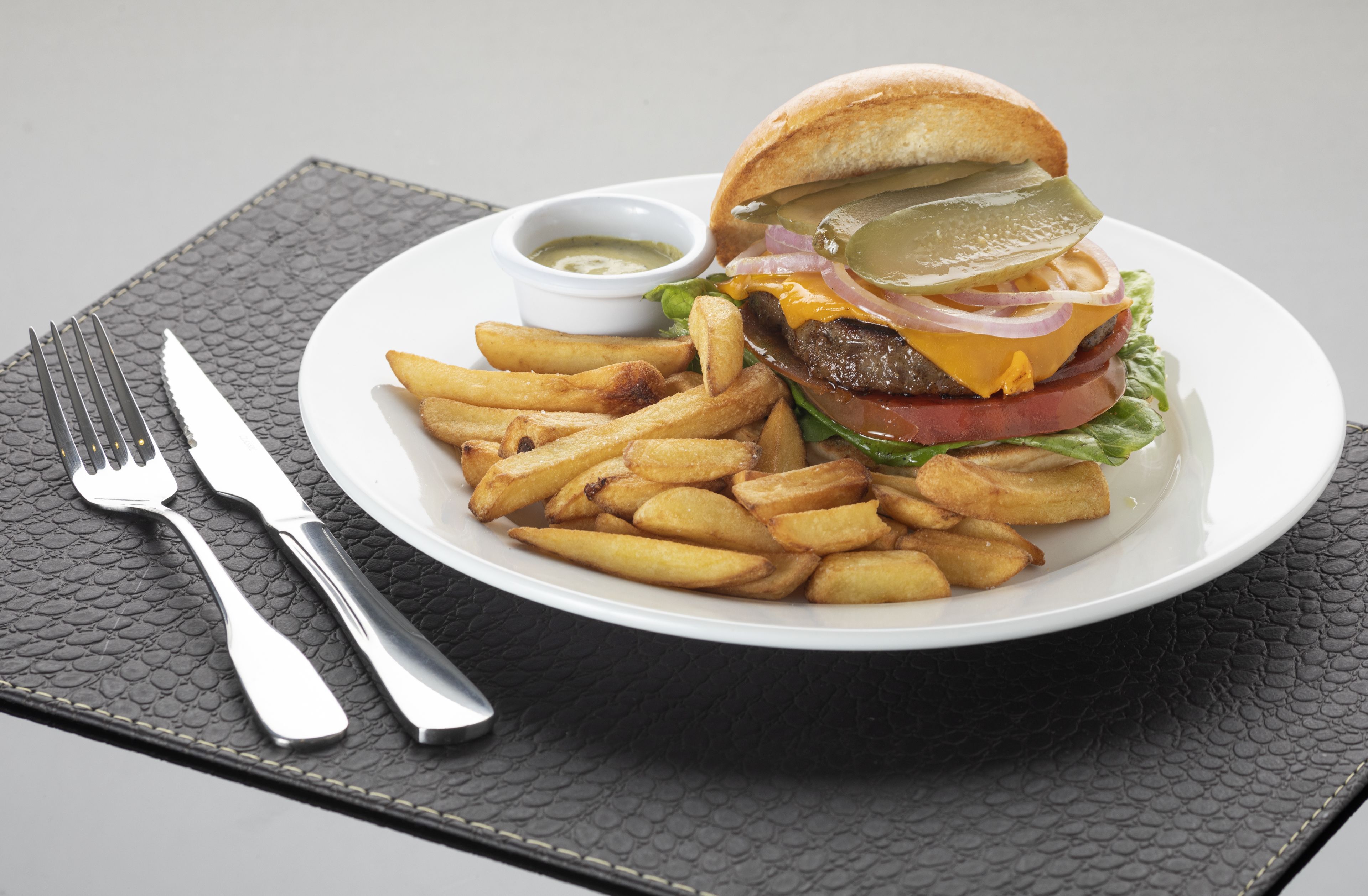 Un plato con una hamburguesa, papas fritas y un recipiente pequeño con salsa sobre un mantel negro con textura.