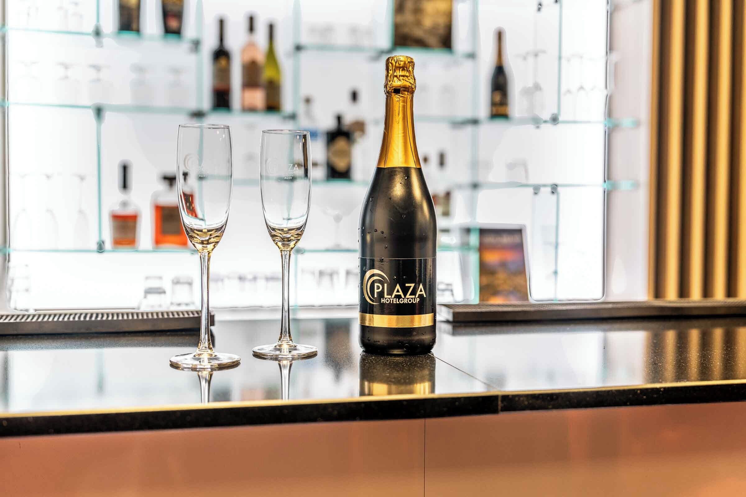 A bar area with a bottle of Plaza Hotel Group champagne and two glasses on a reflective surface.