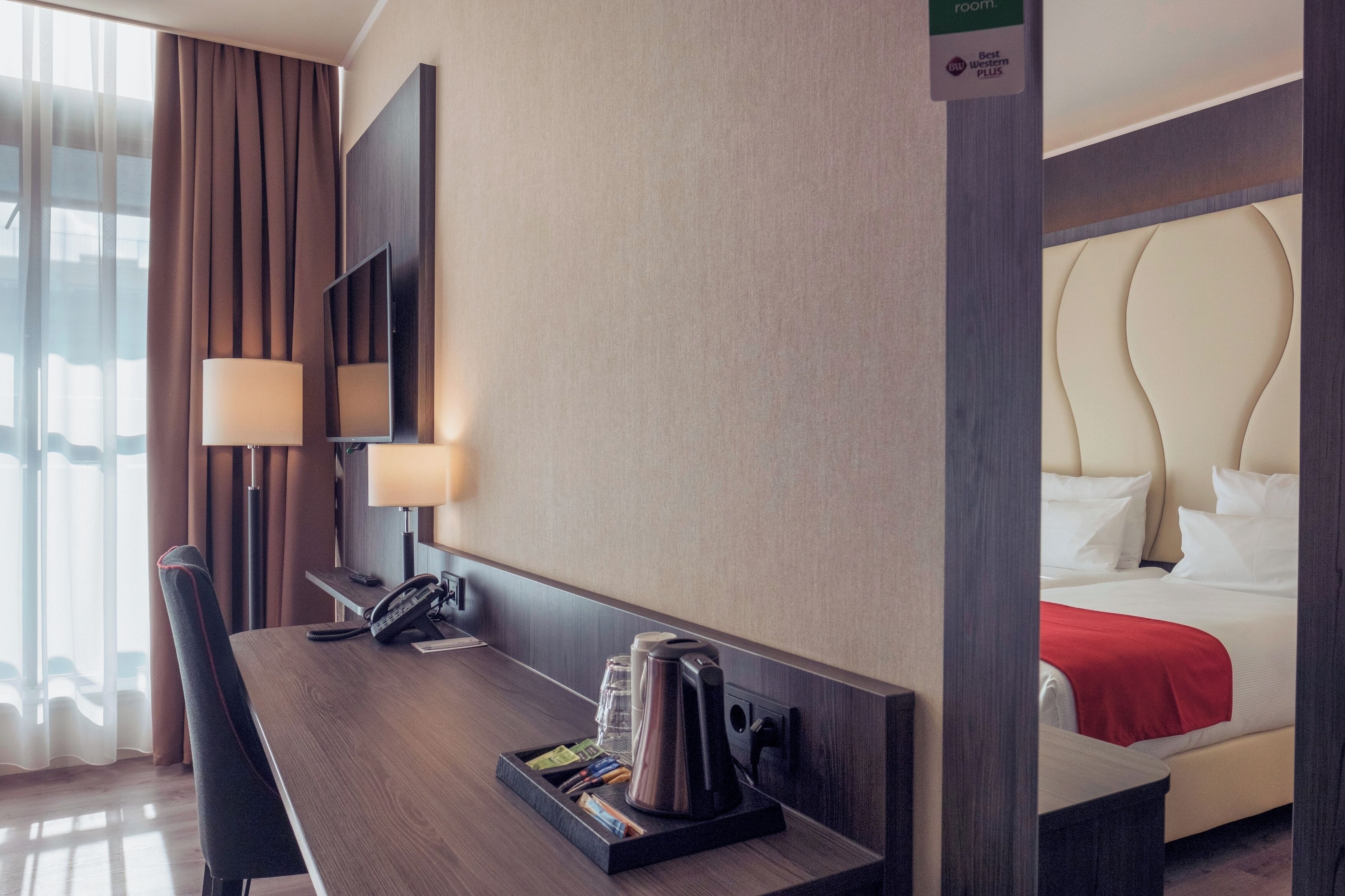 A hotel room with a wooden desk, a telephone, a coffee maker, and a bed with a red blanket.
