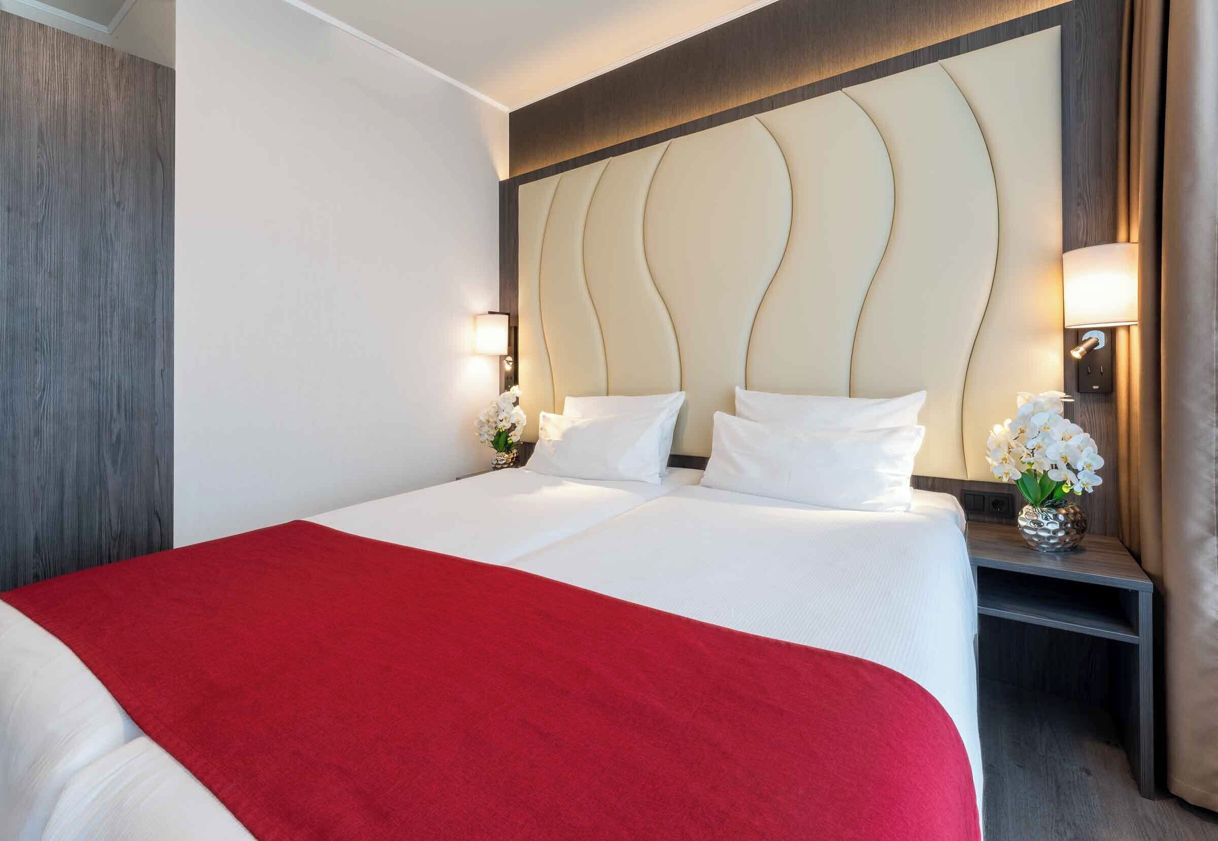 Una habitación de hotel con una cama grande, una manta roja decorativa, un cabecero ornamental, lámparas de noche y un jarrón con flores.