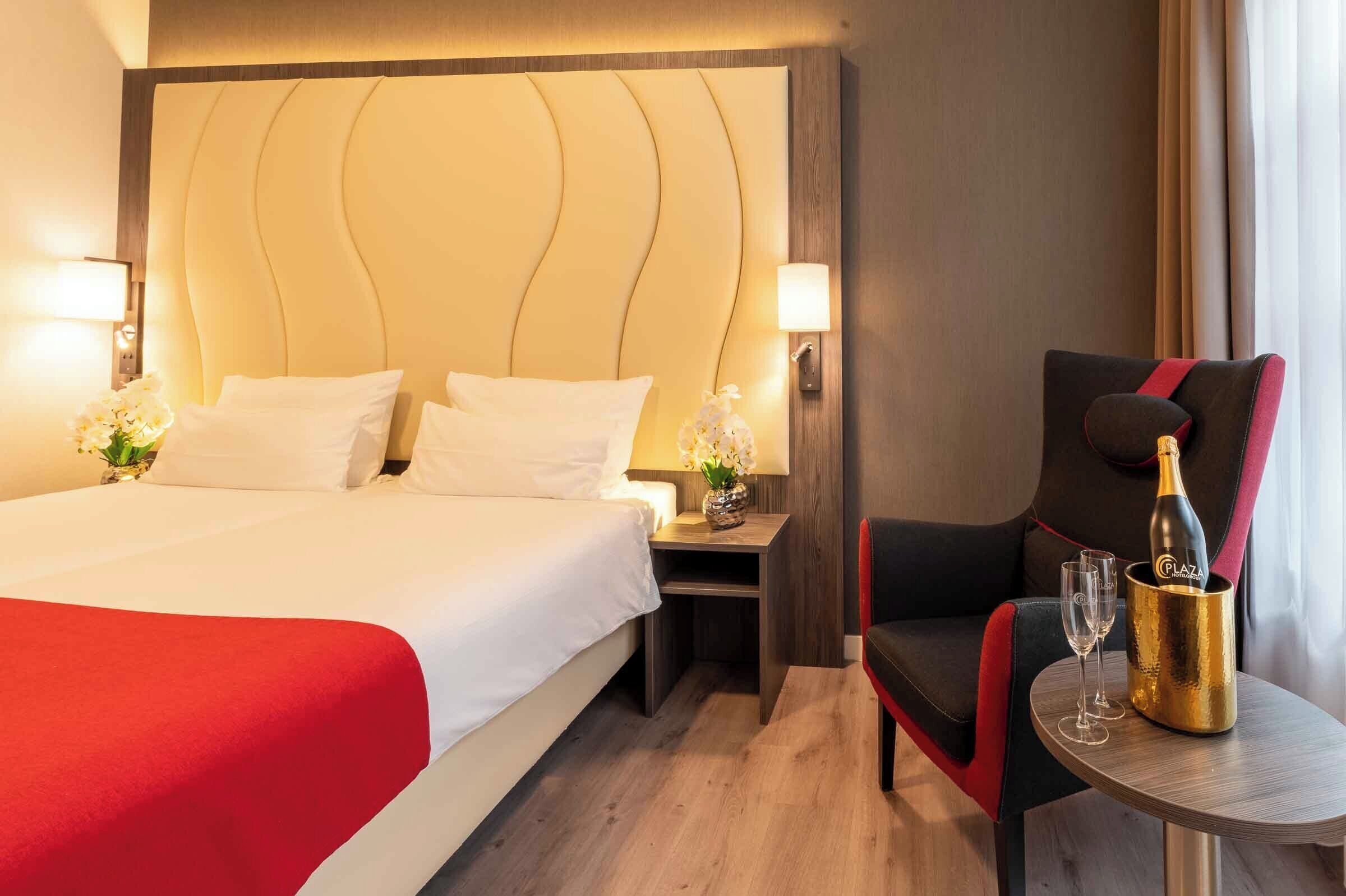 Una habitación de hotel con una cama grande, una mesita de noche con una lámpara, una silla y una mesa con una botella de champán y dos copas.