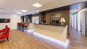 Una moderna zona de recepción de hotel con suelo de madera, un mostrador de recepción y un área de descanso con sillas rojas.